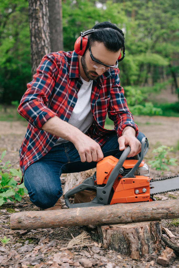 lumberman in plaid shirt and hearing protectors adjusting chainsaw in forest - Photo, Image