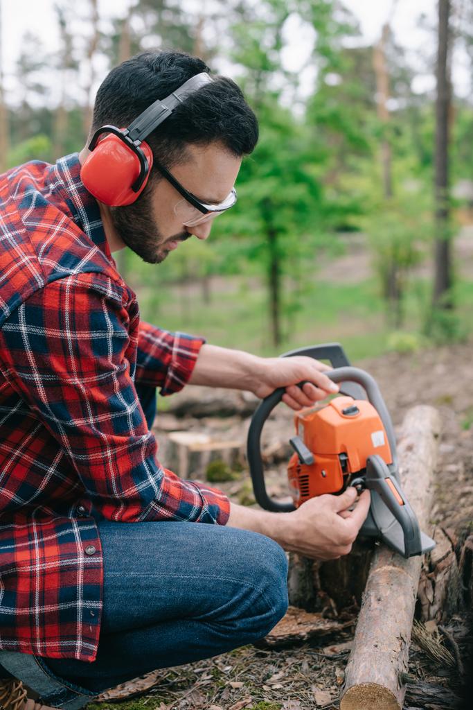 lenhador em fones de ouvido com cancelamento de ruído consertando motosserra na floresta
 - Foto, Imagem
