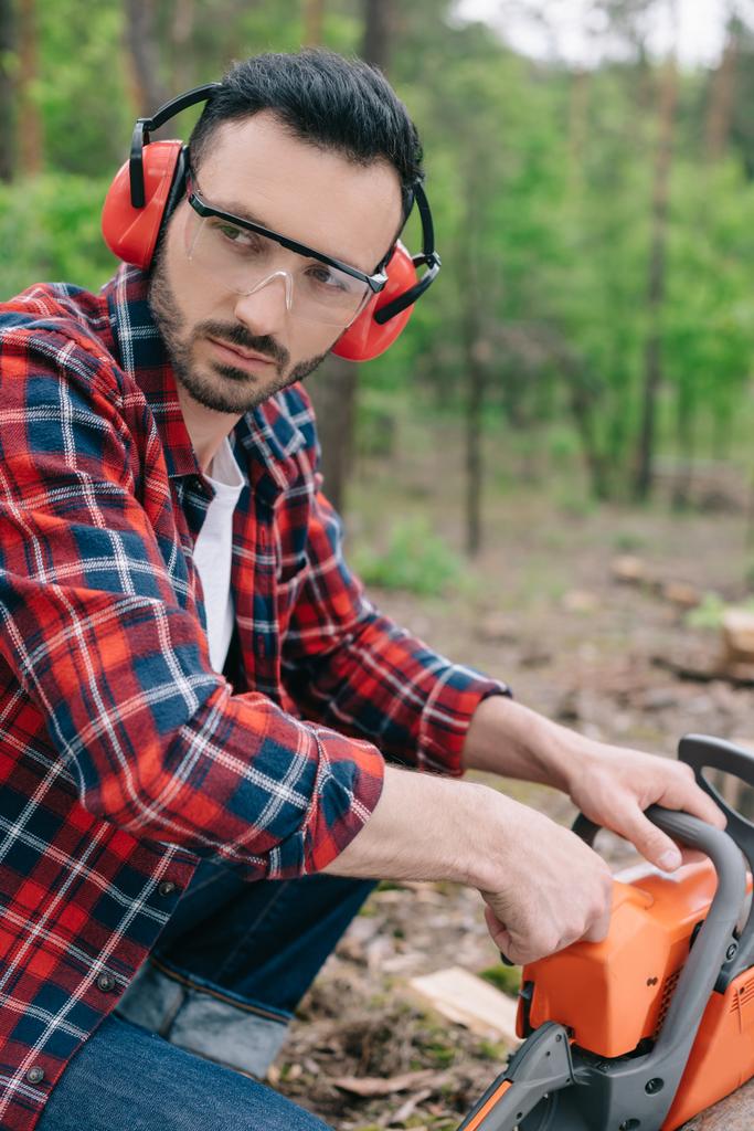 serious lumberjack in earmuffs and hearing protectors looking away in forest - Photo, Image