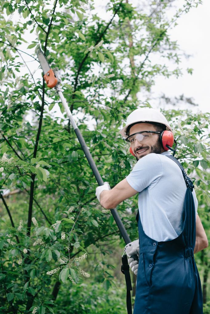 jardinero sonriente en árboles de recorte de casco con sierra telescópica y mirando a la cámara
 - Foto, Imagen