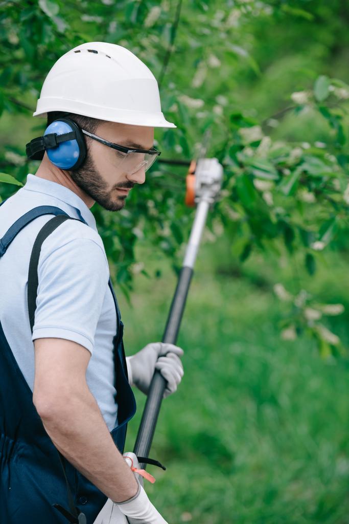 Selektiver Fokus des durchdachten Gärtners mit Helm und Gehörschutz beim Baumschneiden mit Teleskopstangensäge im Garten - Foto, Bild