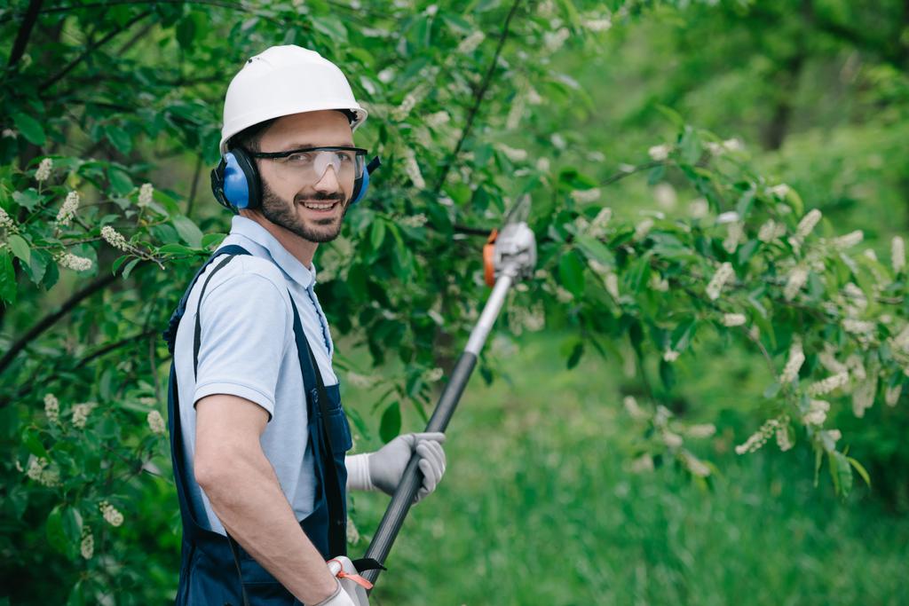 handsome gardener in helmet, protective glasses and noise-canceling headphones holding telescopic pole saw and smiling at camera - Photo, Image