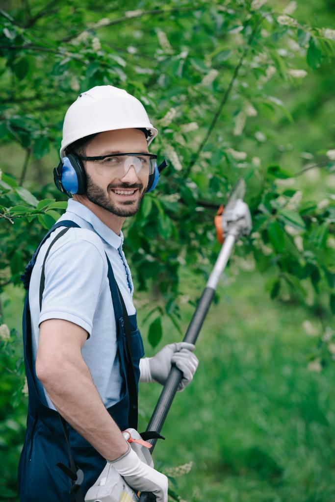 vrolijke tuinman in helm, beschermende bril en ruisonderdrukkende hoofdtelefoon met telescopische paal zaag en glimlachend op de camera - Foto, afbeelding