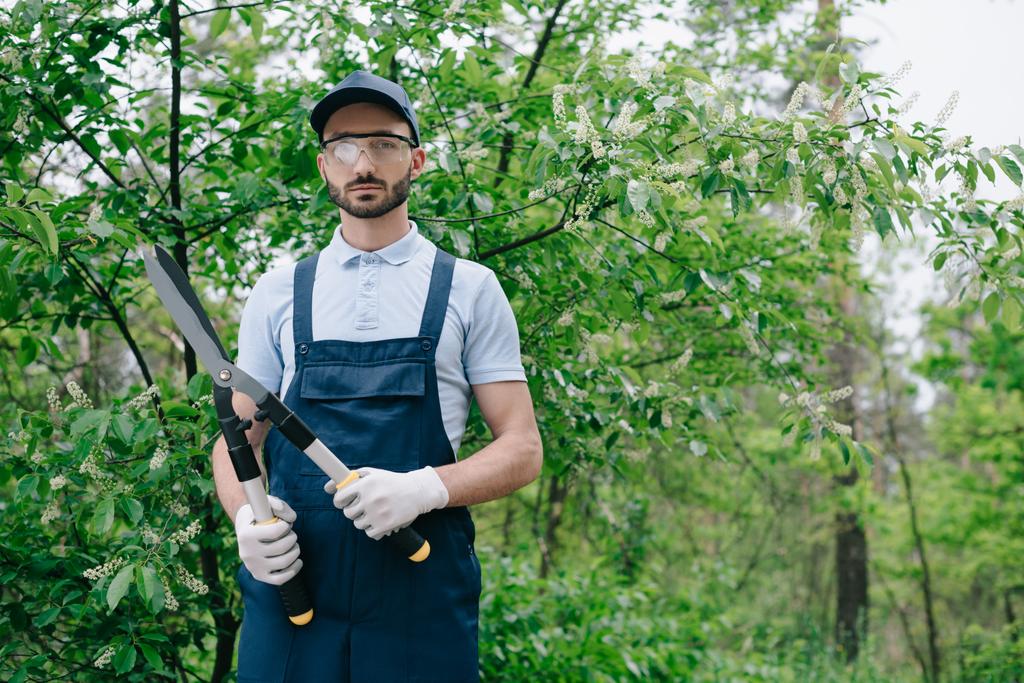 serieuze tuinman in overalls, pet en beschermende glazen houden trimmer en glimlachend op camera - Foto, afbeelding