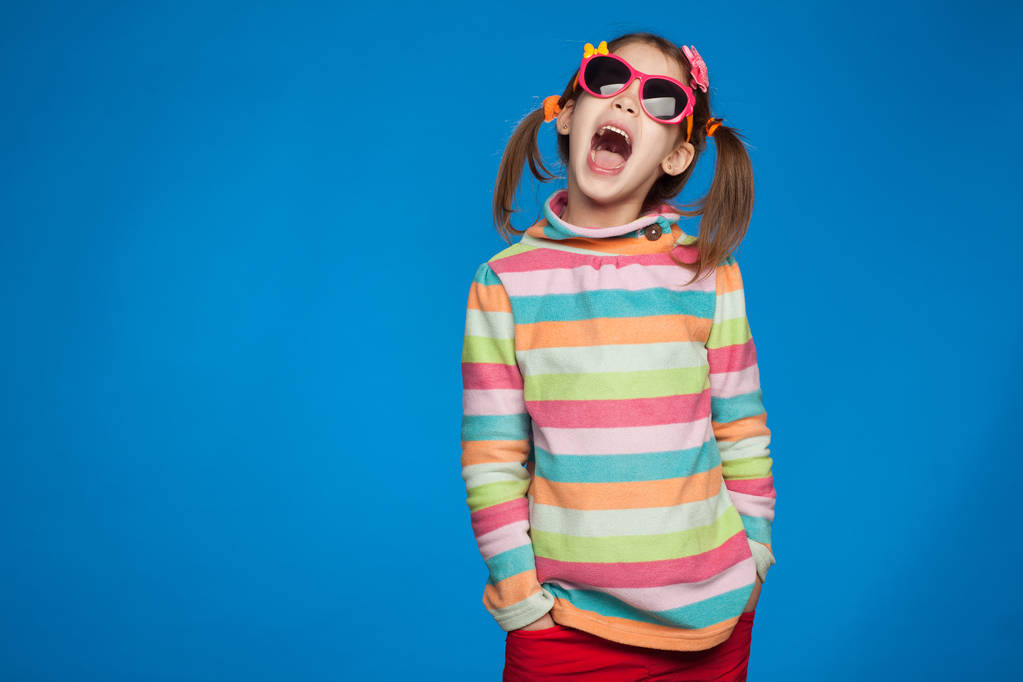 Portrait d'une fille émotionnelle de cinq ans dans un pull rayé et dans des lunettes pour enfants sur fond bleu
 - Photo, image