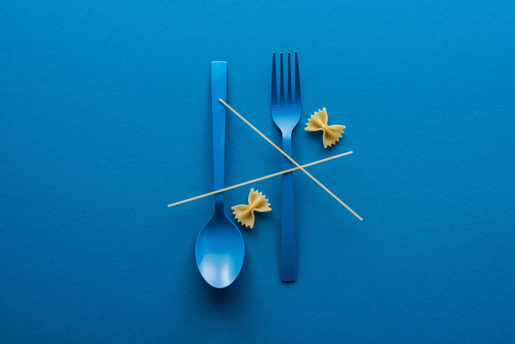 Top uitzicht van spaghetti op blauwe plastic lepel ondersteboven en vork in de buurt van Farfalle pasta op blauwe achtergrond - Foto, afbeelding