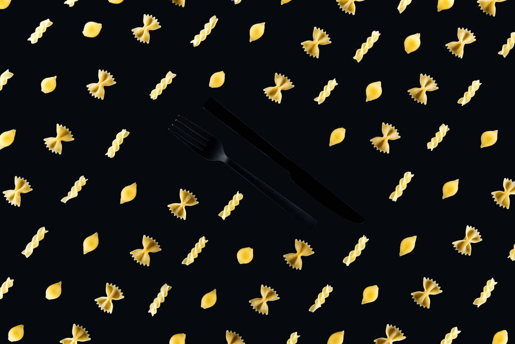 επίπεδη μορφή των διαφόρων ειδών ζυμαρικών με πλαστικό μαύρο πιρούνι και μαχαίρι στη μέση απομονωμένη σε μαύρο - Φωτογραφία, εικόνα