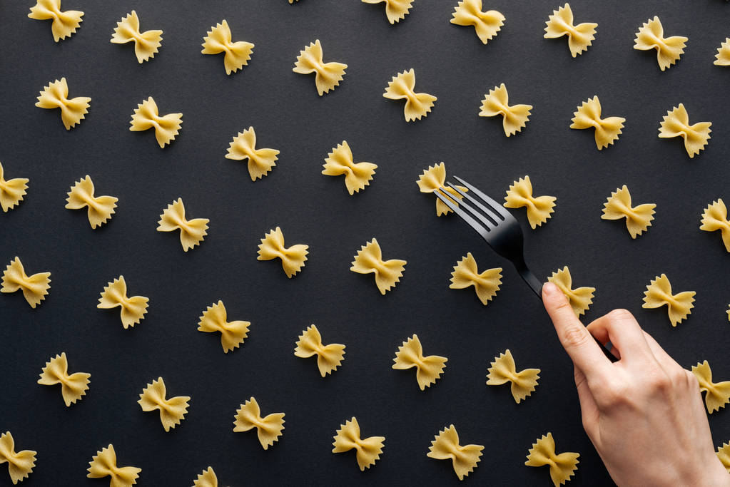 bijgesneden beeld van vrouw die probeert Farfalle pasta te nemen met plastic voorvork geïsoleerd op zwart - Foto, afbeelding
