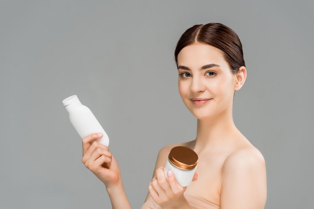 vrolijke brunette vrouw houder met gezichtscrème en fles met body lotion geïsoleerd op grijs - Foto, afbeelding