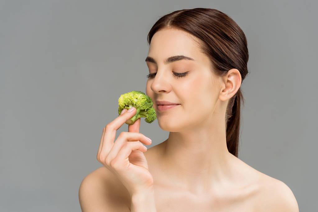 gelukkige naakte vrouw glimlachend terwijl ze groene broccoli geïsoleerd op grijs houdt  - Foto, afbeelding