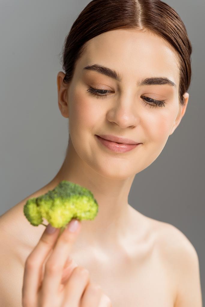 selective focus of happy naked woman smiling while holding green broccoli isolated on grey  - Photo, Image