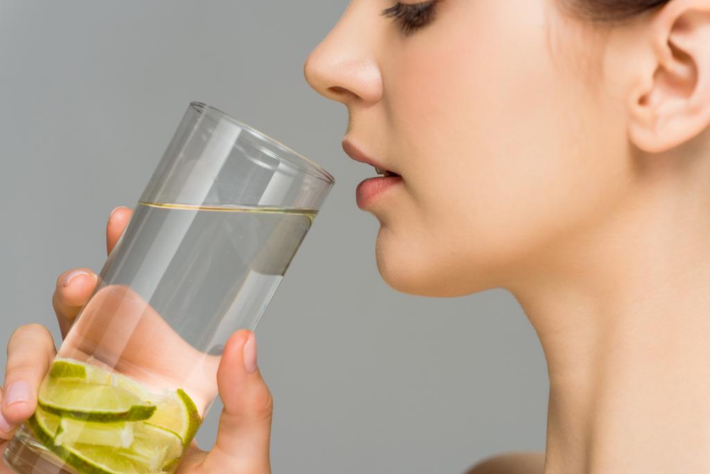 вид сбоку молодой женщины, смотрящей на стакан воды с нарезанной известью, изолированной на сером
  - Фото, изображение