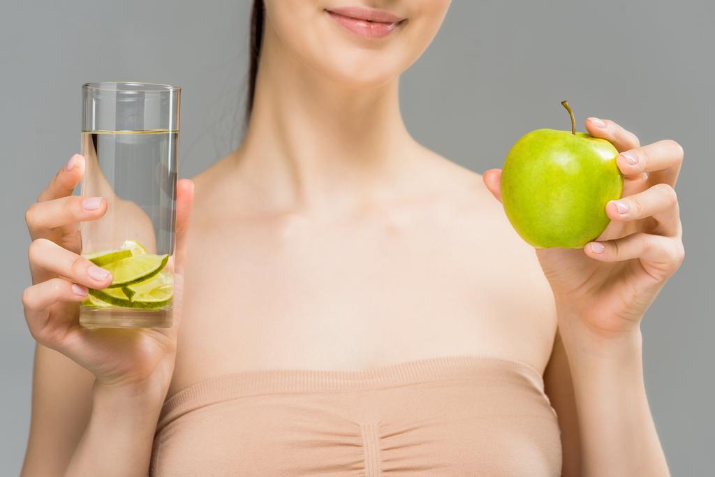 обрізаний вид веселої дівчини, що тримає склянку води з нарізаним лаймом і зеленим яблуком ізольовано на сірому
  - Фото, зображення