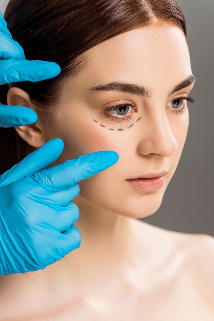 visão recortada de cirurgião plástico em luvas de látex perto de mulher com marcas no rosto isolado em cinza
  - Foto, Imagem