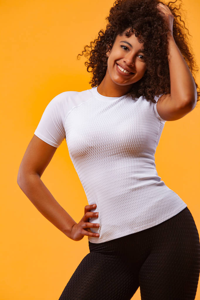 Χαμογελώντας ισχυρή αθλητική γυναίκα με μαύρο δέρμα και σγουρά μαλλιά, κάνει άσκηση σε κίτρινο φόντο φορώντας αθλητικά ρούχα. Άσκηση και κίνητρο αθλητισμού. - Φωτογραφία, εικόνα