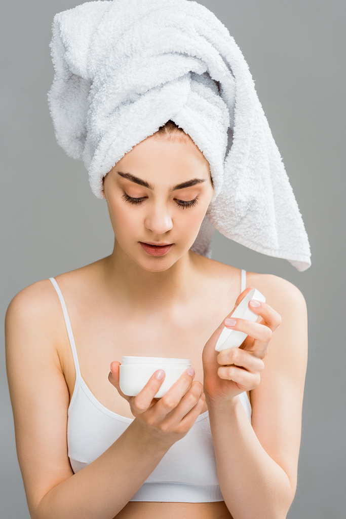 atrakcyjna kobieta z ręcznikiem na głowie patrząc na pojemnik z kremem kosmetycznym białym na szarym  - Zdjęcie, obraz