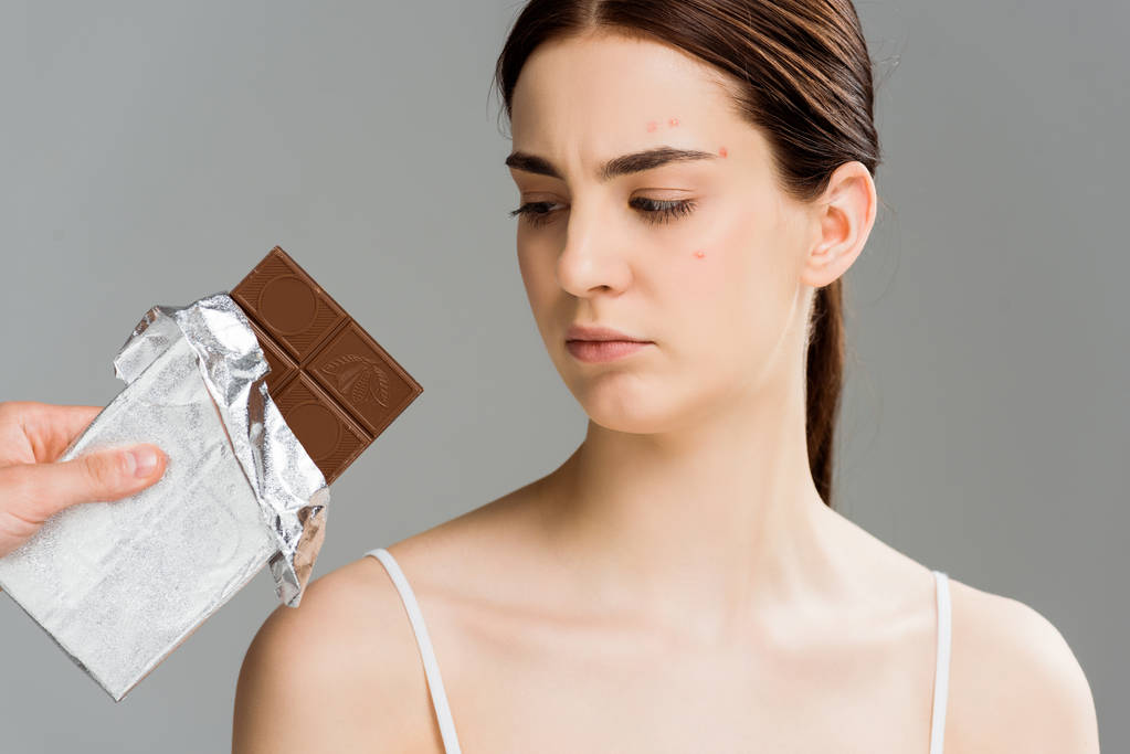περικομμένη όψη του ανθρώπου κρατώντας μπάρα σοκολάτας κοντά μελαχρινή αναστατωμένη γυναίκα με ακμή απομονωμένη σε γκρι  - Φωτογραφία, εικόνα