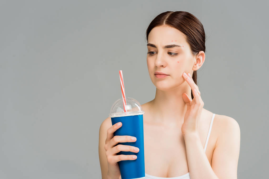 woman with acne on face looking at plastic cup with straw isolated on grey - Photo, Image