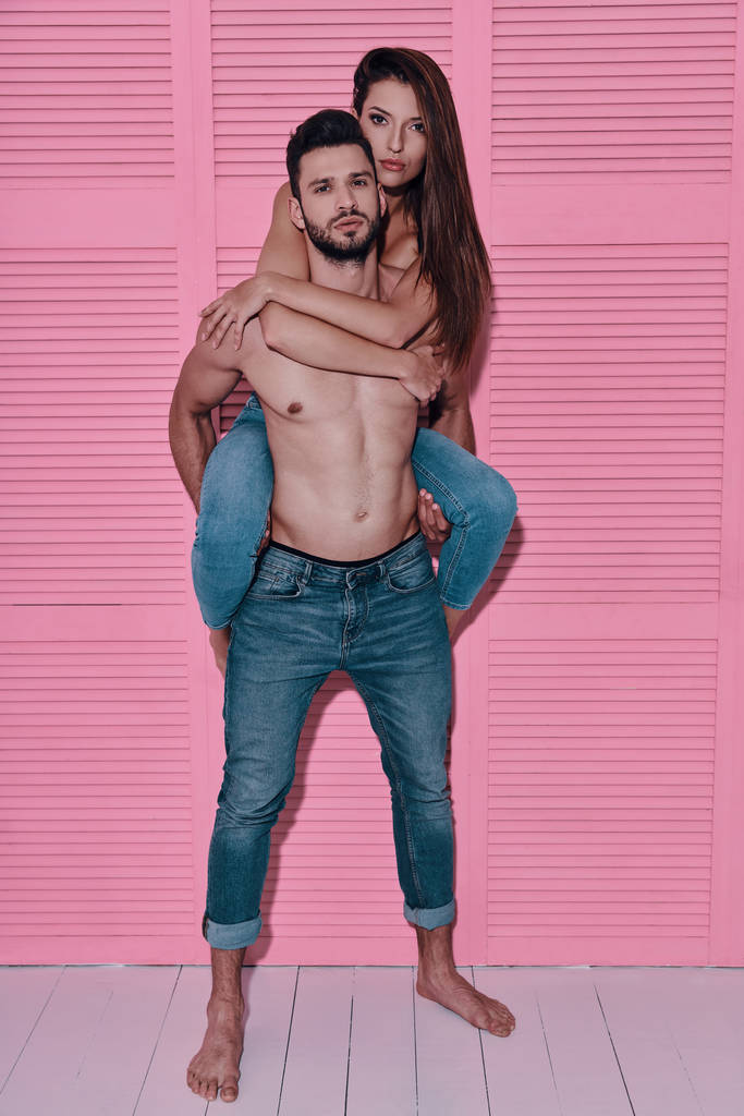 Täysi pituus komea paidaton mies kuljettaa tyttöystävänsä hartioilla, kun seisoo vaaleanpunainen tausta
 - Valokuva, kuva
