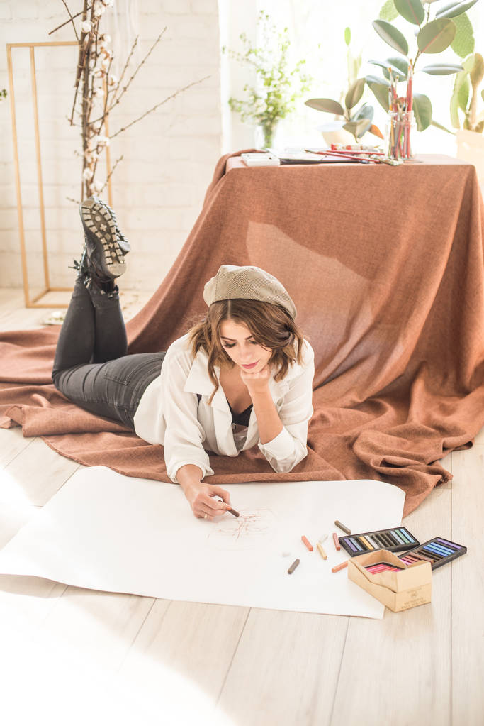 Ελκυστική γυναίκα καλλιτέχνης με σκούρα μαλλιά κάθεται στο πάτωμα σε ένα στούντιο που περιβάλλεται από παστέλ μπογιές χαρτί και μπογιές - Φωτογραφία, εικόνα