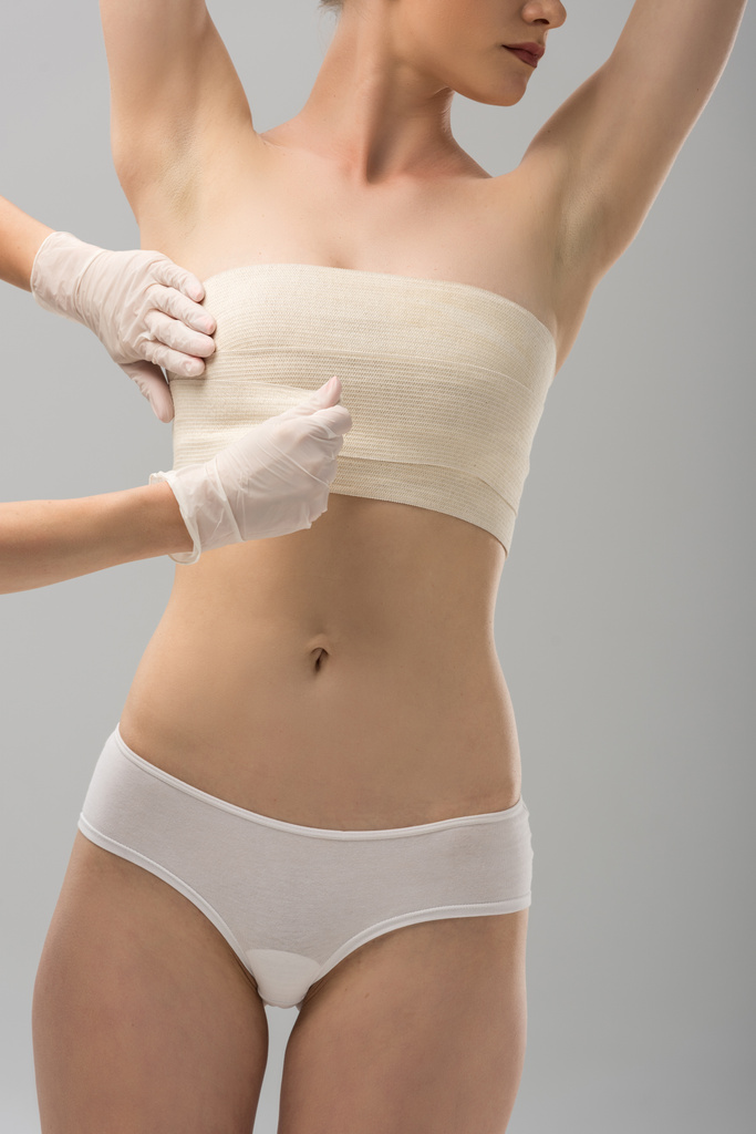 vista parcial del cirujano plástico en guantes de látex y paciente en vendaje mamario aislado en gris
 - Foto, Imagen