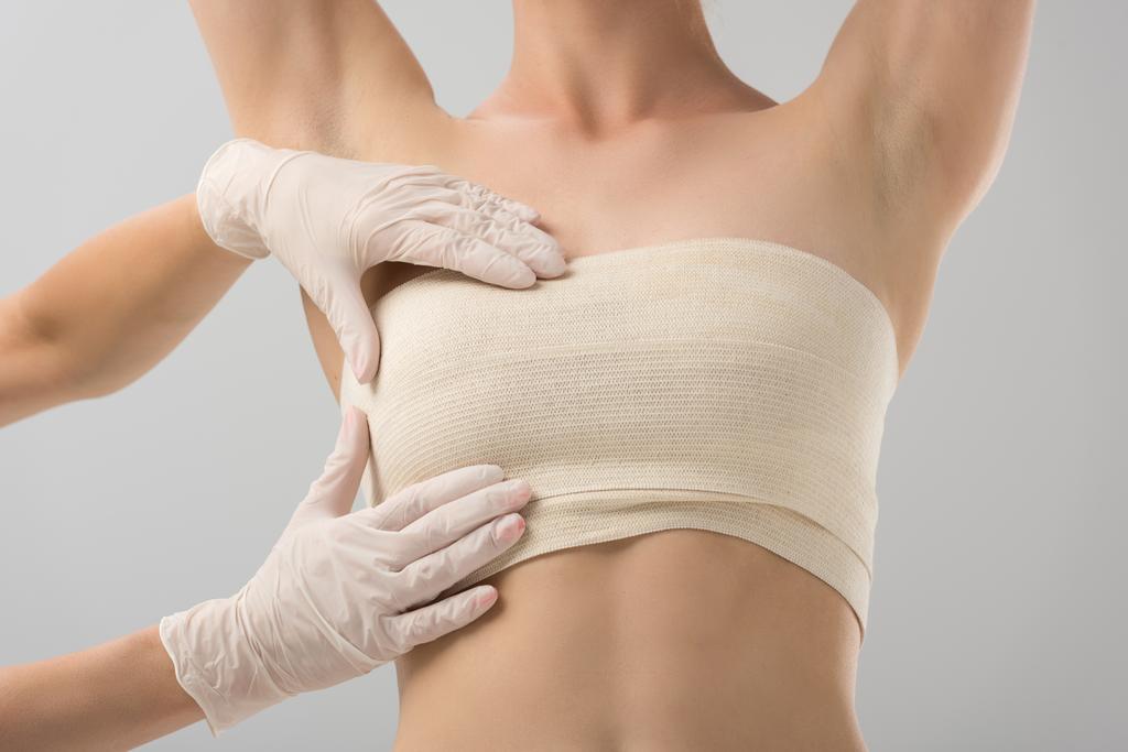 visão parcial do cirurgião plástico em luvas de látex e paciente em curativo de mama isolado em cinza
 - Foto, Imagem