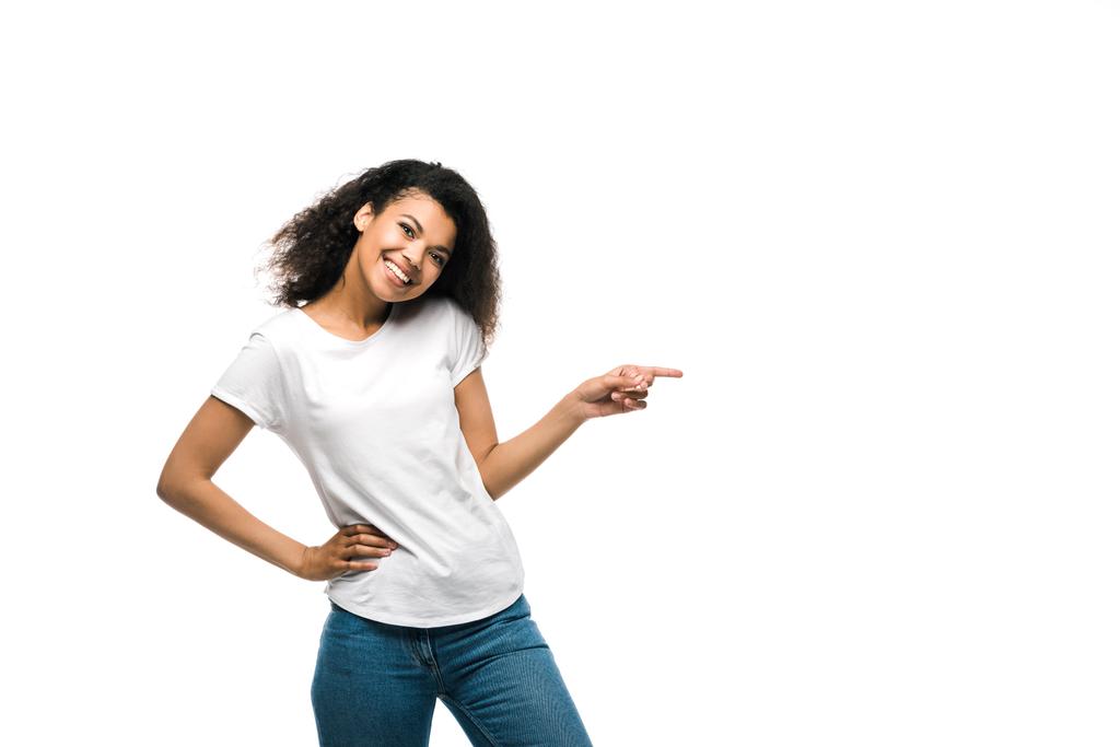ευτυχισμένο αφρικανικό κορίτσι με λευκό t-shirt που σημαδεύει με το δάχτυλο ενώ στέκεται με το χέρι στο ισχίο απομονώνεται σε λευκό  - Φωτογραφία, εικόνα