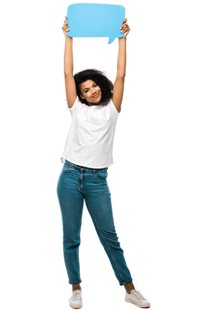 白で隔離された青いジーンズに立っている間、青いスピーチバブルを保持している幸せなアフリカ系アメリカ人の女の子  - 写真・画像