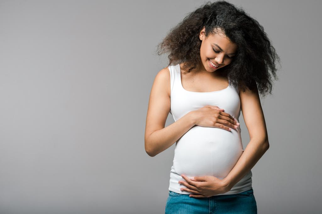 陽気な妊娠中のアフリカ系アメリカ人の女の子が灰色の腹に触れる  - 写真・画像