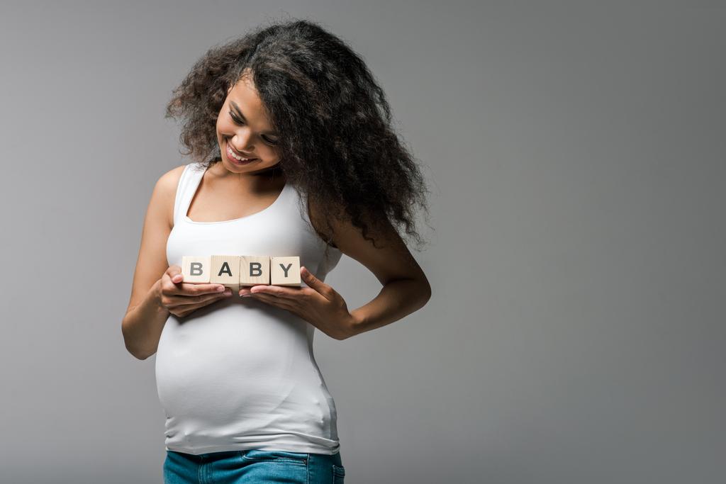 vidám terhes afro-amerikai lány, aki fából készült kockákra baba betűkkel, szürke  - Fotó, kép