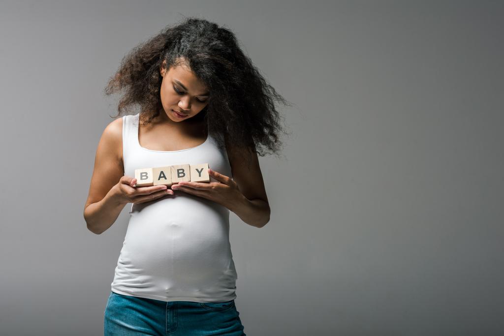 έγκυος αφρικανική αμερικανική γυναίκα στέκεται και κρατώντας ξύλινο κύβους με το μωρό γράμματα σε γκρι  - Φωτογραφία, εικόνα