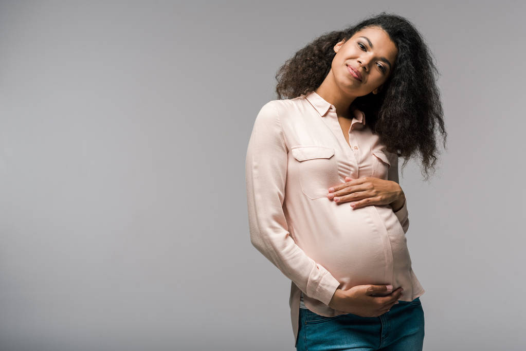 χαμογελαστή νεαρή έγκυος αφρικανική αμερικανική γυναίκα αγγίζοντας την κοιλιά, ενώ στέκεται στο γκρι  - Φωτογραφία, εικόνα
