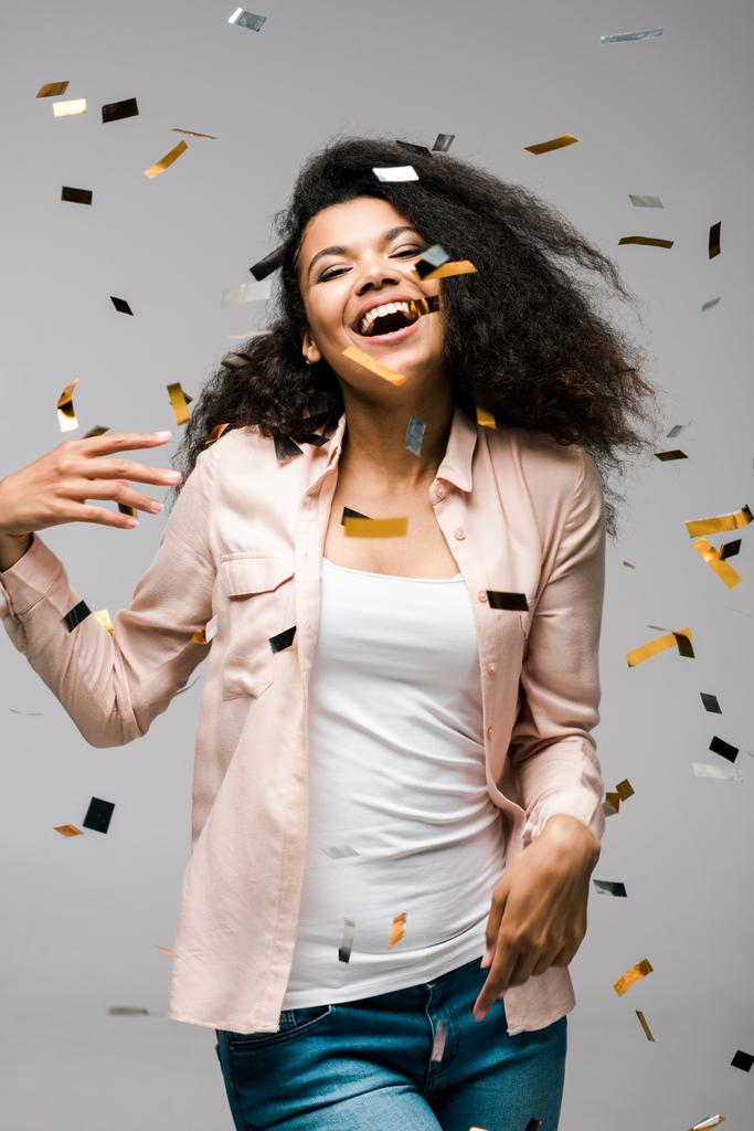 Gelukkig African American Girl glimlachend in de buurt van glanzende confetti en gebaren op grijs  - Foto, afbeelding