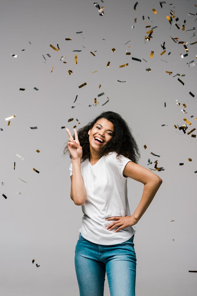young african american woman smiling near shiny confetti while standing with hand on hip and showing peace sign on grey  - Photo, Image