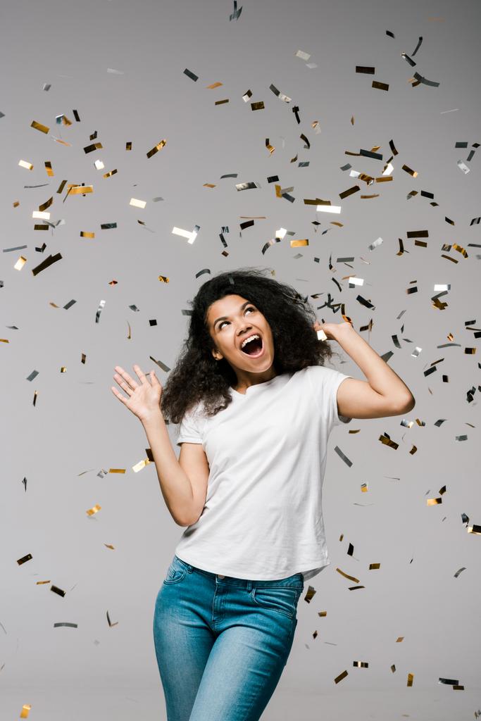 mladá Africká Američanka usmívajíje se a dívá se na padající konfety, když stojí na šedé  - Fotografie, Obrázek
