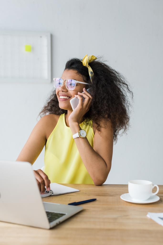 dizüstü bilgisayar yakınında otururken akıllı telefon konuşurken gözlük mutlu Afrikalı Amerikalı kadının seçici odak  - Fotoğraf, Görsel