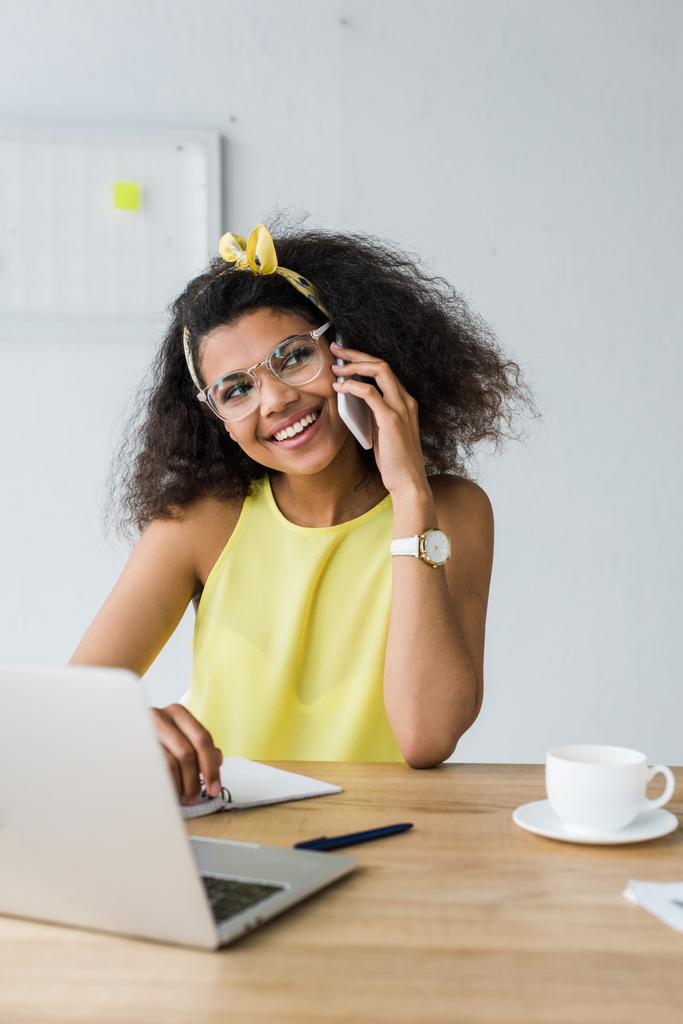 enfoque selectivo de la alegre mujer de negocios afroamericana en gafas hablando en el teléfono inteligente mientras está sentado cerca de la computadora portátil
  - Foto, imagen