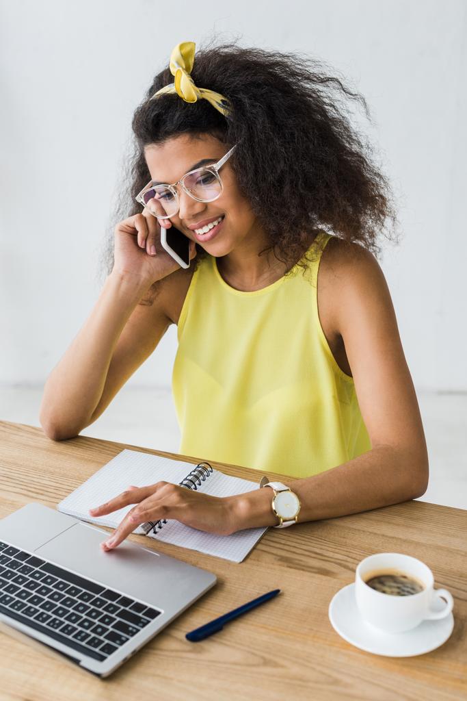 счастливая африканская американка в очках с помощью ноутбука, разговаривая на смартфоне возле чашки кофе
 - Фото, изображение