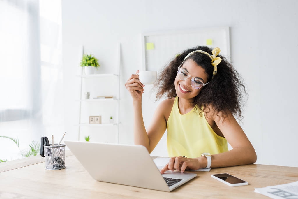 щаслива афроамериканська бізнес-леді в окулярах дивиться на ноутбук, тримаючи чашку
  - Фото, зображення