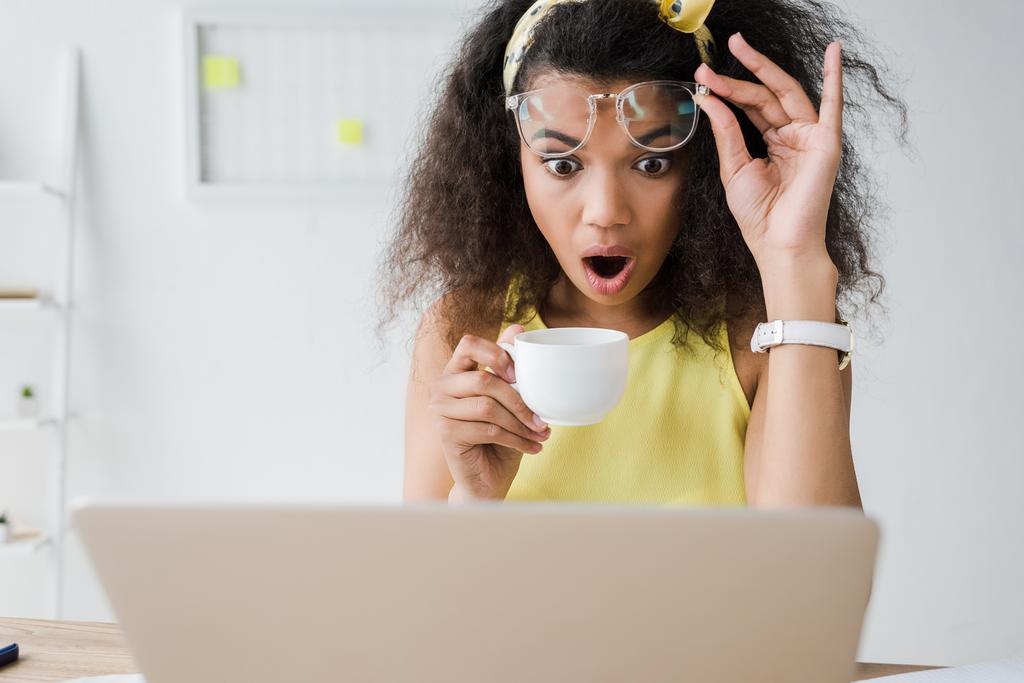 foyer sélectif de choqué afro-américain fille toucher des lunettes tout en regardant ordinateur portable et tenant tasse de café
  - Photo, image