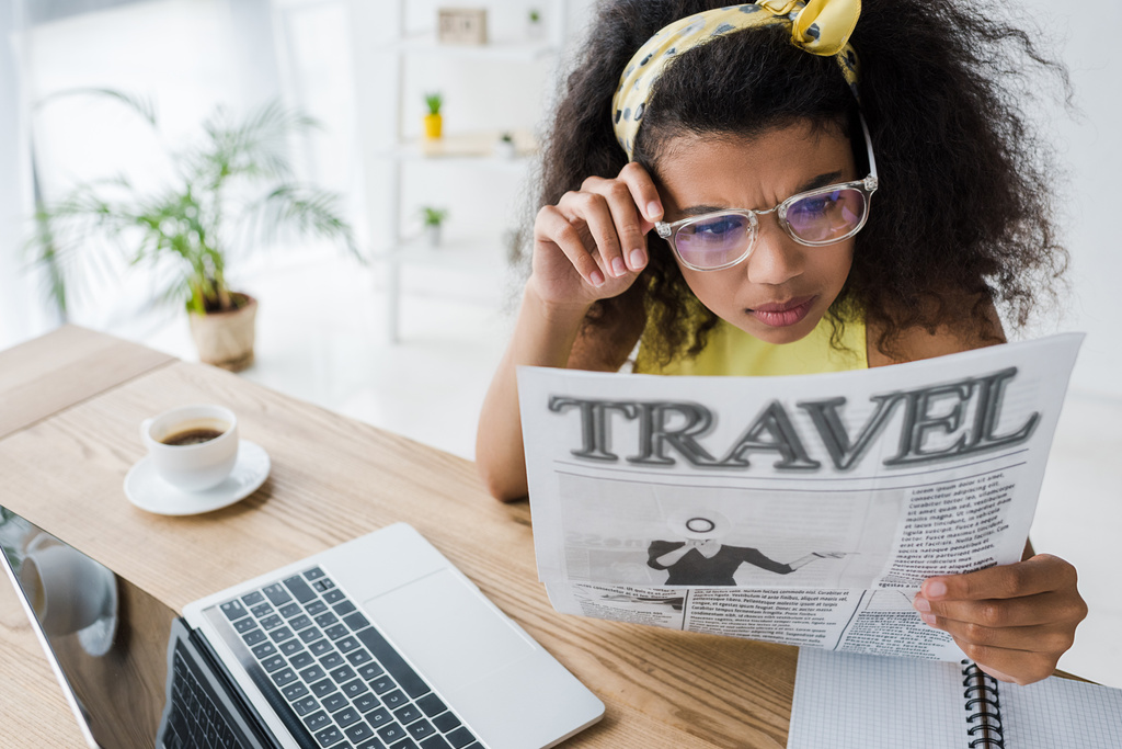 attraente donna afro-americana in occhiali lettura giornale di viaggio vicino al computer portatile
  - Foto, immagini