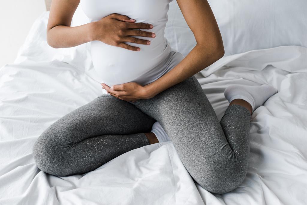 abgeschnittene Ansicht einer schwangeren Afroamerikanerin, die ihren Bauch berührt, während sie auf dem Bett sitzt  - Foto, Bild