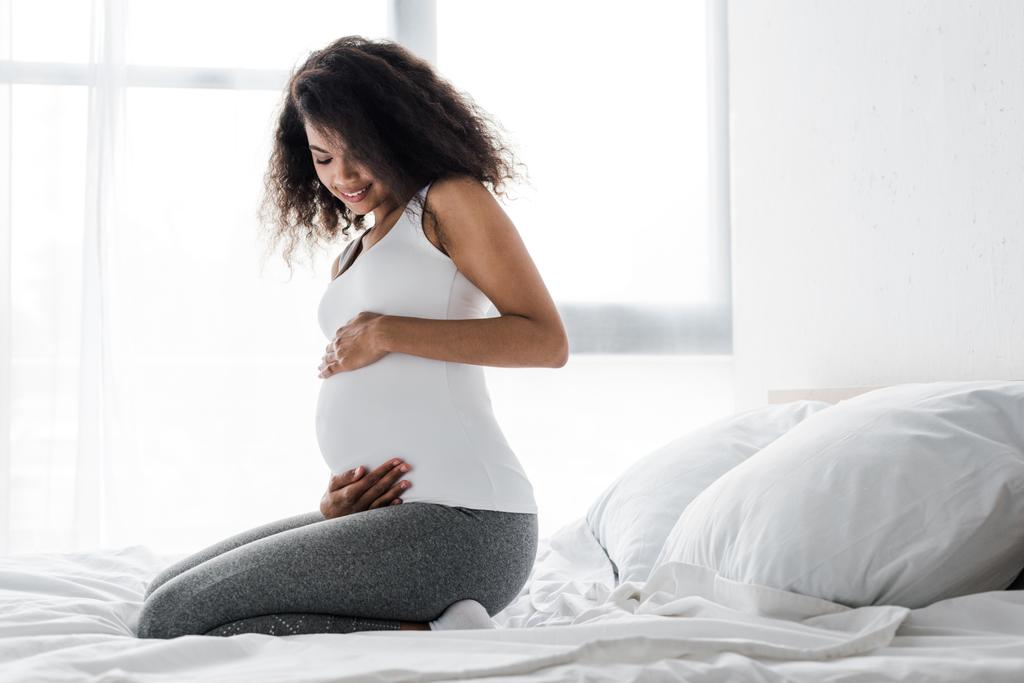 mosolyogva terhes afro-amerikai nő megható hasa, miközben ül az ágyban  - Fotó, kép