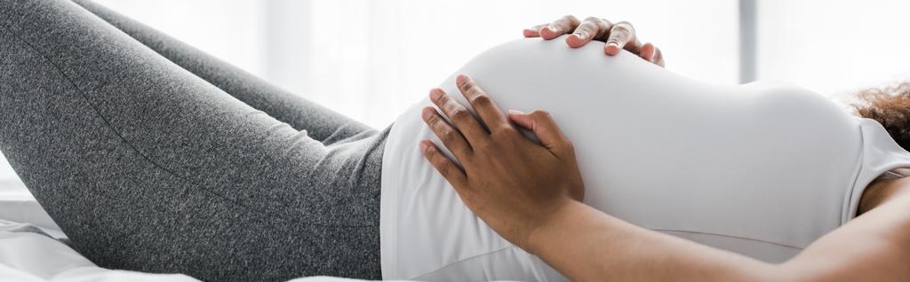 Panoramaaufnahme einer schwangeren Afroamerikanerin, die ihren Bauch berührt, während sie auf dem Bett liegt  - Foto, Bild