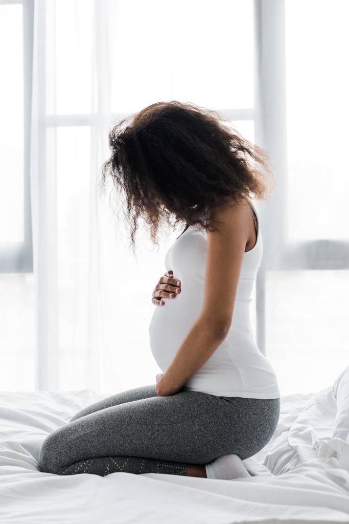 σγουρά έγκυος αφρικανική Αμερικάνικη γυναίκα που αγγίζει την κοιλιά ενώ κάθεται στο κρεβάτι  - Φωτογραφία, εικόνα