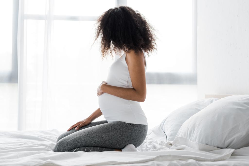 έγκυος αφρικανική αμερικανική γυναίκα αγγίζοντας την κοιλιά, ενώ κάθεται στο κρεβάτι  - Φωτογραφία, εικόνα