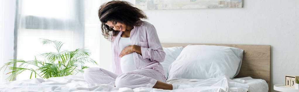 魅力的な妊娠中のアフリカ系アメリカ人女性のパノラマショットは、腹を見て、ベッドに座って  - 写真・画像