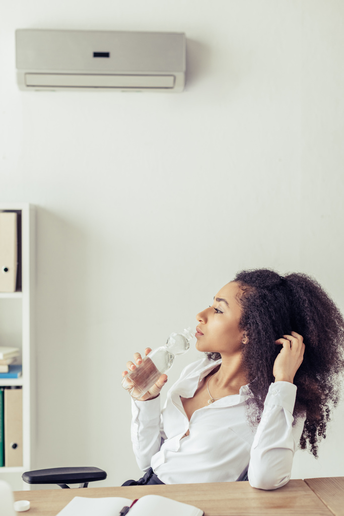pensativa mujer de negocios afroamericana beber agua mientras está sentado bajo el aire acondicionado en la oficina
 - Foto, imagen