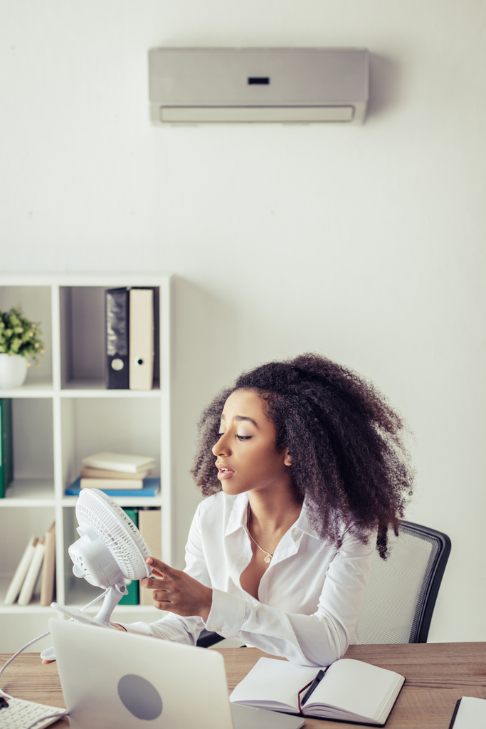 Afrikanisch-amerikanische Geschäftsfrau hält blasenden elektrischen Ventilator, während sie am Arbeitsplatz unter Klimaanlage sitzt - Foto, Bild