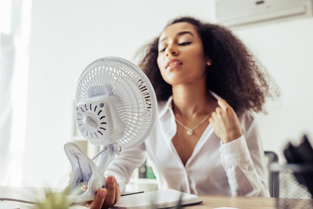selektivní zaměření atraktivní africké americké obchodní ženy držící elektrický ventilátor při sezení na pracovišti  - Fotografie, Obrázek
